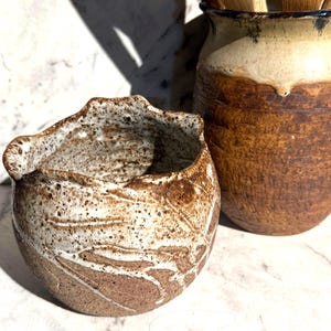 Pode incluir: Duas peças de cerâmica artesanais. Uma tigela pequena e redonda com uma superfície texturizada e salpicada em tons de castanho e branco. Ao lado, um porta-utensílios cilíndrico mais alto com uma paleta de cores semelhante. Ambas as peças têm uma estética rústica e artesanal.