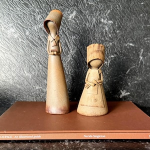 May include: Two ceramic figurines of figures in long dresses and bonnets. The taller figure is standing, and the shorter figure is seated. Both are in a neutral colour palette with a matte finish. The figurines are on a brown book.