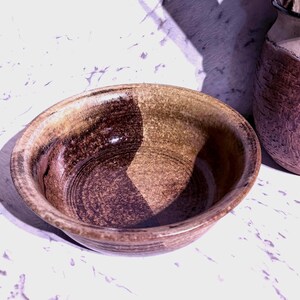May include: A round ceramic bowl with a speckled, earthy brown and tan glaze. The interior features a distinct dark brown circular pattern contrasted against a lighter tan base, with a smooth, curved rim and a sturdy, thick-walled construction.