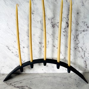 May include: A black arched candle holder with five tapered slots, each holding a tall, slender, light yellow beeswax candle. The curved base is made of a matte black material, providing a stable foundation for the five vertical candles.
