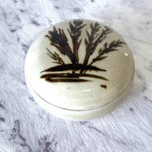 May include: A small, round, ceramic trinket box with a lid. The box is off-white with a dark brown tree design on the lid. The design is a stylized depiction of a tree with branches and leaves. The box is likely handmade.