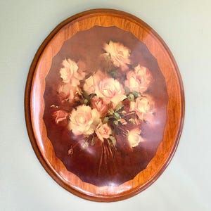 Può includere: Vassoio ovale in legno con un dipinto floreale di rose rosa chiaro e foglie verdi. Il dipinto è incorniciato in un ovale marrone scuro, bordato da un bordo in legno più chiaro. Un oggetto decorativo.