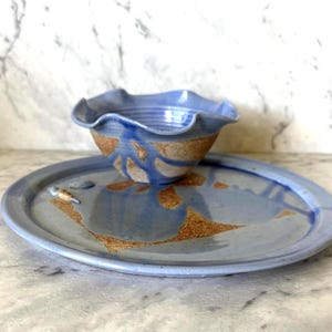 May include: A ceramic bowl with a ruffled edge and a matching flat plate. Both pieces feature a glossy periwinkle blue glaze applied over a textured, speckled tan clay base, creating a distinct contrast between the smooth blue and raw earth tones.