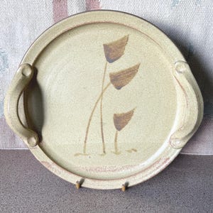 May include: A round, beige ceramic serving tray with two curved handles. The tray features a hand-painted design of three brown abstract flowers. The rim has a darker brown accent. The tray is supported by two small, gold-coloured stands.