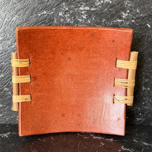May include: A square, slightly curved terracotta-coloured ceramic dish with a speckled finish. The dish features two vertical wooden dowels on the left and right sides, each secured to the ceramic body with three sets of light-coloured woven rattan.