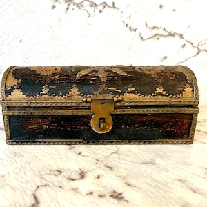 Op de afbeelding: Een kleine, antieke houten kist met een gebogen bovenkant. De kist is donkerbruin met messing accenten en een messing sluiting. De bovenkant en randen zijn versierd met ingewikkelde bloempatronen. De kist wordt waarschijnlijk gebruikt voor het opbergen van sieraden of kleine souvenirs.