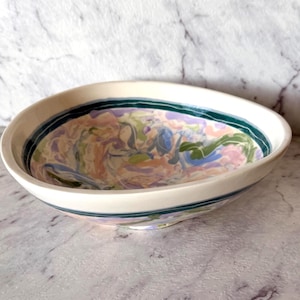 May include: A shallow ceramic bowl with a cream-coloured exterior and a wide, dark green painted rim. The interior features a marbled pattern with swirls of pastel pink, lavender, light blue, and sage green colours across the basin surface.
