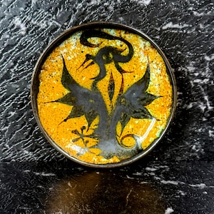 Puede incluir: Un plato decorativo redondo con un fondo amarillo vibrante y un diseño floral oscuro y estilizado. El borde del plato es de color marrón oscuro y la obra de arte presenta acentos negros y verdes. El plato es probablemente de cerámica o un material similar.