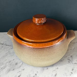 May include: A ceramic casserole dish with a lid. The dish is a light beige color with a brown rim and lid. The lid has a small, round handle. The dish has two handles on either side.