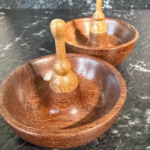 May include: Two wooden bowls with attached pestles. The bowls are a rich brown colour, and the pestles are lighter in tone. The bowls have a smooth, polished finish. The pestles are attached to the centre of the bowls.