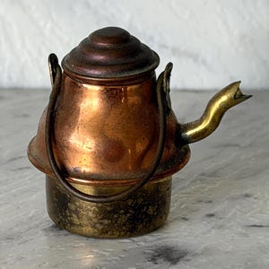 May include: A miniature metal kettle featuring a polished copper upper body with a dark, rounded lid and a brass-toned lower base. The kettle includes a curved brass spout and a thin, dark metal swing handle attached to the sides of the body.