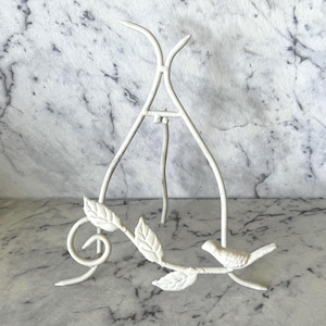 May include: A white metal display stand featuring a decorative bird and leaf branch. The stand has a tripod base and a curved top. The bird is perched on a branch with leaves. The stand is painted white.