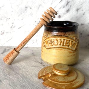 May include: A ceramic honey pot with a yellow and brown speckled glaze, featuring the word HONEY embossed on the front. The pot includes a matching circular lid with a small notch and a long wooden honey dipper resting against the rim.
