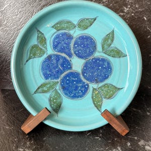 May include: A turquoise ceramic plate with a hand-painted design of blue plums and green leaves.