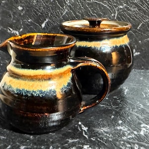 May include: A handmade ceramic creamer and covered jar set. The pottery features a dark brown base with a band of yellow and blue glaze around the middle. The creamer has a handle and spout. The jar has a lid with a small knob.