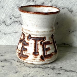 May include: A white ceramic mug with a flared rim and a wide base, featuring the name PETER embossed in large, dark brown capital letters around the centre. The piece has a speckled texture and a dark brown glazed rim and lettering.