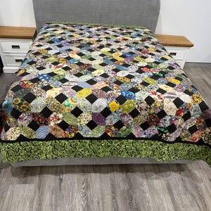 Op de afbeelding: Een handgemaakte quilt met een geometrisch patroon van kleurrijke bloemen en stoffen met patronen, tegen zwarte vierkanten. De quilt heeft een groene rand met bladeren en wordt getoond op een bed met een grijs hoofdbord en houten nachtkastjes.