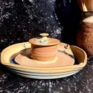 May include: Handmade ceramic serving dish with a small lidded container in the centre. The dish is a light tan colour with a darker brown and blue accent. The lidded container is a similar colour and design. A rustic kitchenware item.