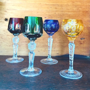 May include: Four vintage crystal wine glasses in vibrant colours. The glasses have clear, decorative stems and bases. The bowls are ruby red, emerald green, sapphire blue, and amber yellow. The yellow glass features a starburst design.