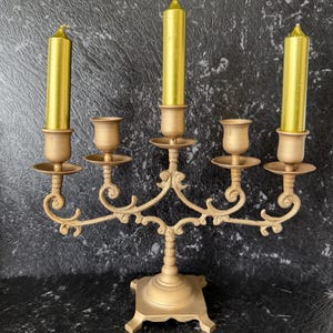 May include: A gold-coloured metal candelabra with five candle holders and a square base. Three metallic gold taper candles are inserted into the centre and two outer holders, while the two remaining holders are empty. The frame features scrollwork.