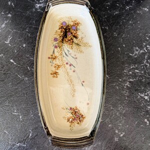 Puede incluir: Fuente de cerámica color crema con forma rectangular y bordes redondeados. Presenta un diseño floral en tonos marrón, morado y verde. El borde está delineado con un borde marrón oscuro. Tiene asas en cada extremo.