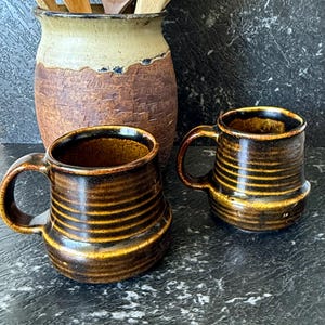 May include: Two brown ceramic mugs with horizontal yellow stripes and curved handles. A larger, textured brown and tan ceramic jar with wooden spoons is in the background. The mugs have a rustic, handmade appearance. The mugs are suitable for tea or coffee.