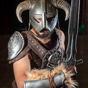 May include: A detailed Skyrim warrior cosplay. The costume features a silver helmet with curved horns, a brown leather and metal chest piece, and a large sword. The arm guards have fur trim.