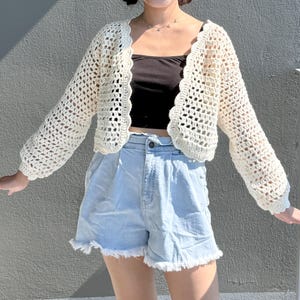 Op de afbeelding: Crèmekleurig, open geweven gehaakt vest met lange mouwen. Het vest heeft een geschulpte rand en wordt gedragen over een zwarte top en lichtblauwe spijkerbroekshorts. De outfit is casual en geschikt voor warm weer.