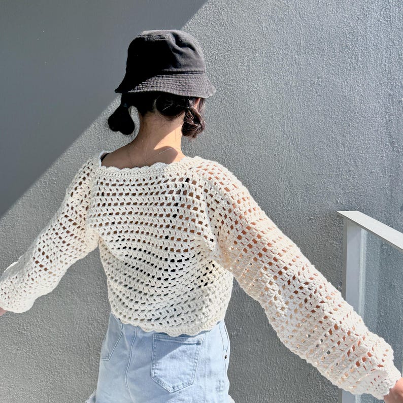 May include: Cream-colored crochet top with long sleeves and a square neckline. The open weave design of the top is visible. A black bucket hat is worn. The top is paired with light blue denim shorts.