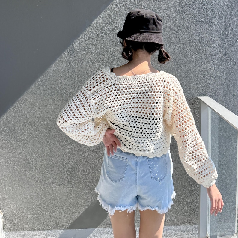 May include: Cream-colored, open-weave crochet sweater with long sleeves. The sweater is paired with light blue denim shorts with frayed edges. A black bucket hat is worn.
