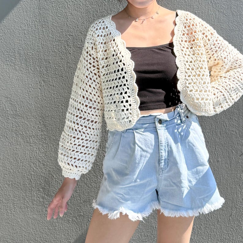 May include: Cream-colored, open-weave crochet cardigan with scalloped edges. The cropped sweater has long sleeves and is paired with a black top and light blue denim shorts with frayed hems. The outfit is set against a gray wall.