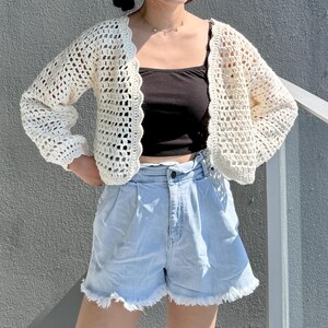 May include: A cream-colored, open-front crochet cardigan with long sleeves and a scalloped edge. The cardigan is paired with a black tank top and light blue denim shorts with a frayed hem. The outfit is casual and summery.