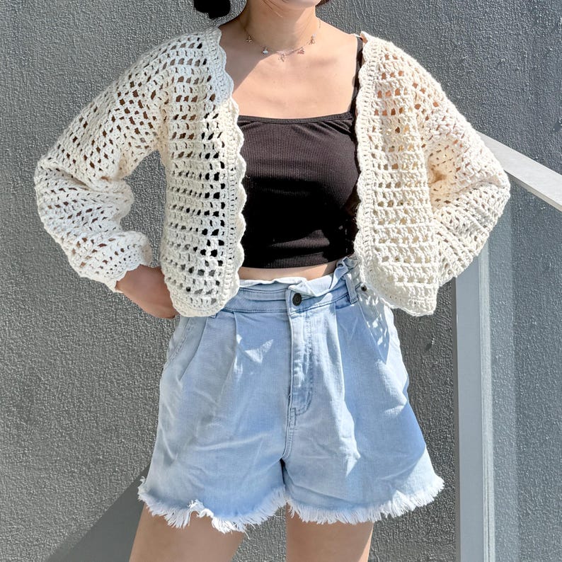 May include: A cream-colored, open-front crochet cardigan with long sleeves and a scalloped edge. The cardigan is paired with a black tank top and light blue denim shorts with a frayed hem. The outfit is casual and summery.