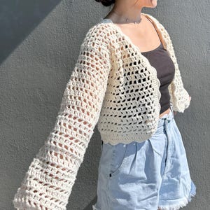 May include: Cream-colored, open-weave crochet cardigan with long sleeves. The cardigan has a scalloped edge and a loose, airy design. The garment is paired with a dark top and light blue denim shorts. The background is a solid gray wall.