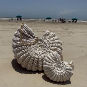 May include: Two off-white crochet bags shaped like nautilus shells, featuring a spiraled, ribbed texture and gold-tone metal chain straps. The bags are crafted from thick yarn, with one larger bag and one smaller matching bag positioned together.