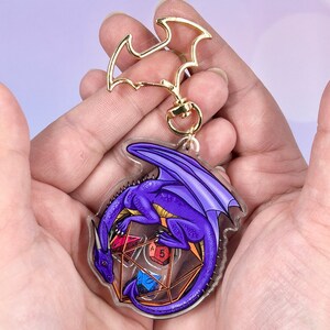 May include: Purple dragon keychain with a clear acrylic charm featuring a dragon curled around a set of dice. The dragon is a deep purple with gold accents. The dice are red, blue, green, and yellow.