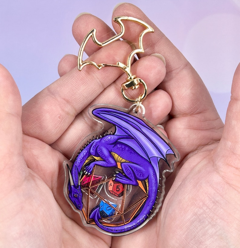 May include: Purple dragon keychain with a clear acrylic charm featuring a dragon curled around a set of dice. The dragon is a deep purple with gold accents. The dice are red, blue, green, and yellow.