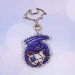May include: A purple dragon curled around a clear, multi-sided die with gold accents. The dragon has a gold-colored eye and a detailed design. The die has a blue, red, and yellow die inside. The charm is attached to a gold-colored key chain.