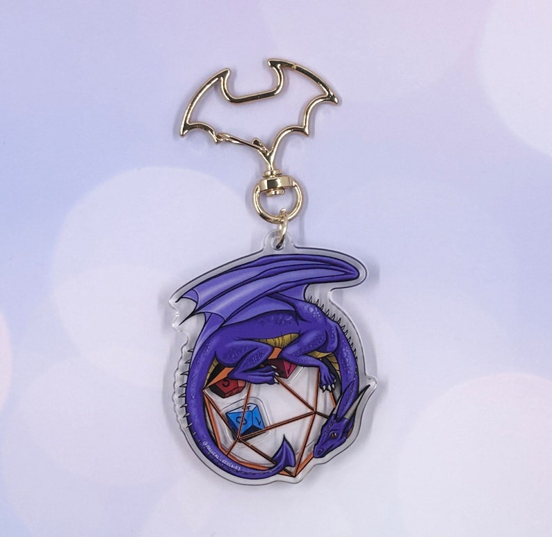 May include: A purple dragon curled around a clear, multi-sided die with gold accents. The dragon has a gold-colored eye and a detailed design. The die has a blue, red, and yellow die inside. The charm is attached to a gold-colored key chain.