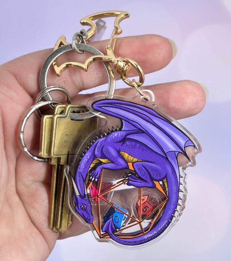May include: Purple dragon keychain with a clear acrylic charm featuring a dragon curled around a set of dice. The keychain is attached to a gold metal key ring.