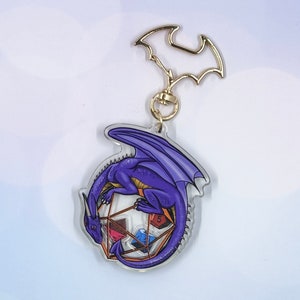 May include: Purple dragon keychain with a clear acrylic background. The dragon is curled around a clear, multi-sided die with colorful dice inside.
