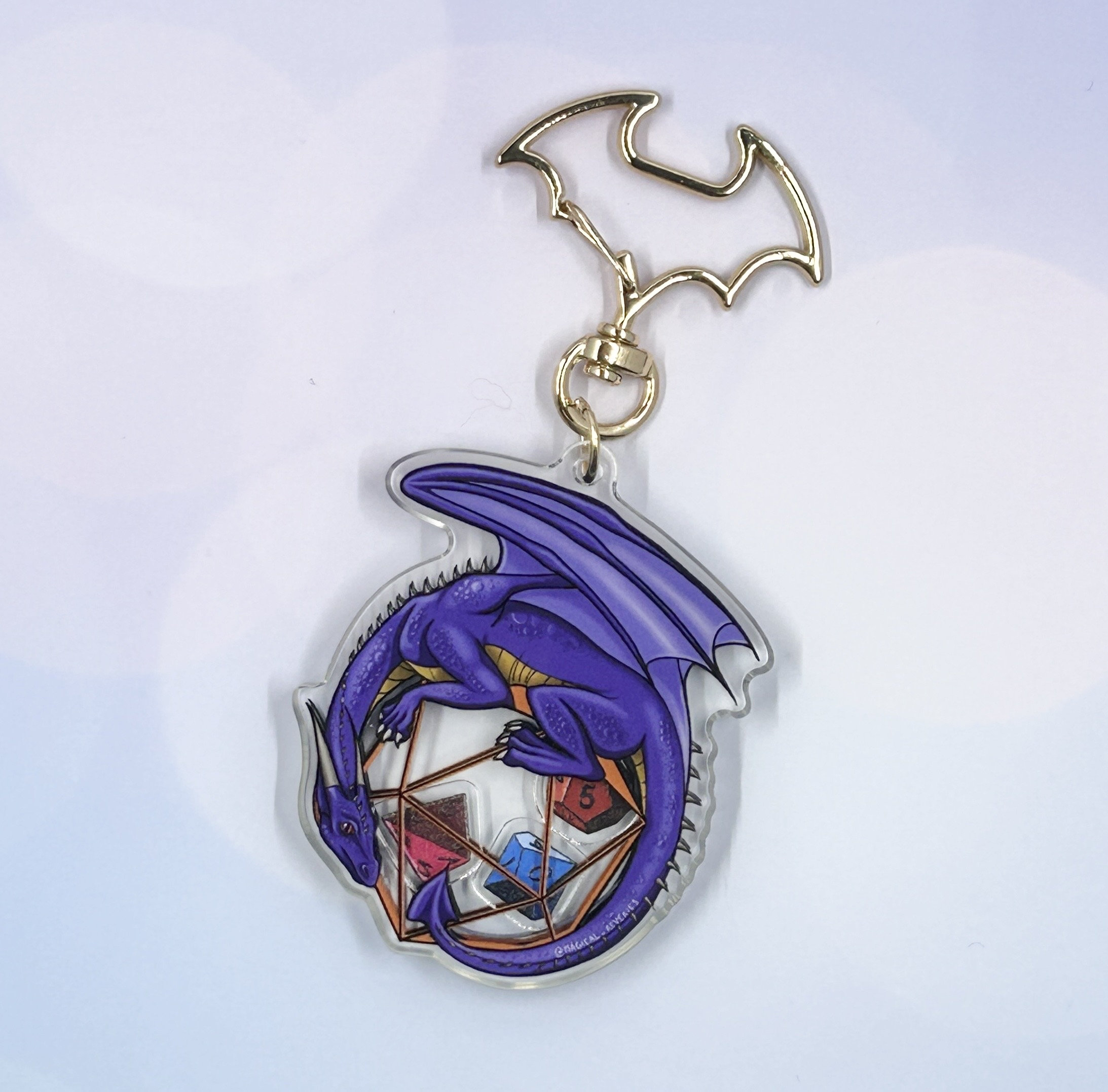 May include: Purple dragon keychain with a clear acrylic background. The dragon is curled around a clear, multi-sided die with colorful dice inside.