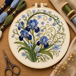 Puede incluir: Un bastidor de bordado que contiene un panel de tela con flores de iris azules y pequeñas flores azul claro cosidas a mano, rodeadas de enredaderas verdes en espiral. El bastidor se acompaña de madejas de hilo y tijeras de metal.