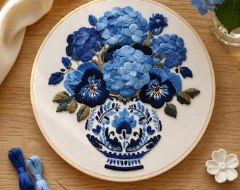 May include: A circular wooden embroidery hoop containing a fabric panel with a hand-stitched design of blue hydrangea and pansy flowers arranged in a decorative blue and white patterned vase, featuring shades of navy, sky blue, and olive green.