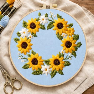 Peut inclure: Un tambour à broder circulaire en bois contenant un tissu bleu clair avec une couronne brodée à la main composée de tournesols jaunes, de marguerites blanches et de feuilles vertes. Le motif est centré, entouré de fils et de ciseaux.
