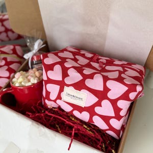 May include: A gift box containing a red mug filled with marshmallows, a red and pink heart-patterned zippered pouch, and other treats. The pouch has a white label that reads "CottonellaDreams". The box is lined with red shredded paper.