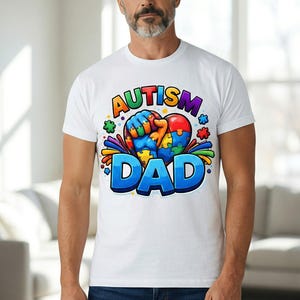 May include: White t-shirt with the words "AUTISM DAD" in a colorful, cartoon-style font. The design includes a heart-shaped puzzle and a fist. The shirt is worn by a person wearing blue jeans.