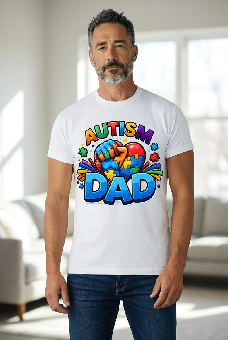 May include: White t-shirt with the words "AUTISM DAD" in a colorful, cartoon-style font. The design includes a heart-shaped puzzle and a fist. The shirt is worn by a person wearing blue jeans.