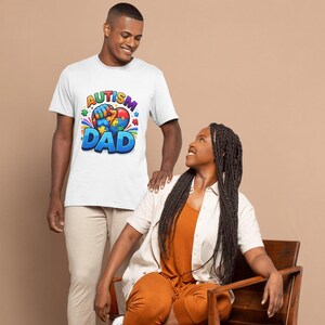May include: A white t-shirt with the text "Autism Dad" in blue, yellow, and red. The shirt features a colorful graphic of puzzle pieces and a heart. A man is wearing the shirt, standing next to a woman.