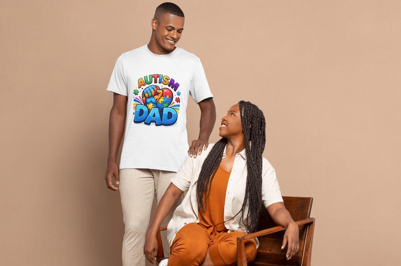 May include: A white t-shirt with the text "Autism Dad" in blue, yellow, and red. The shirt features a colorful graphic of puzzle pieces and a heart. A man is wearing the shirt, standing next to a woman.